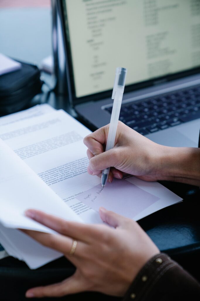 Close-up of hands writing on documents with a laptop, indicating productivity and professionalism.